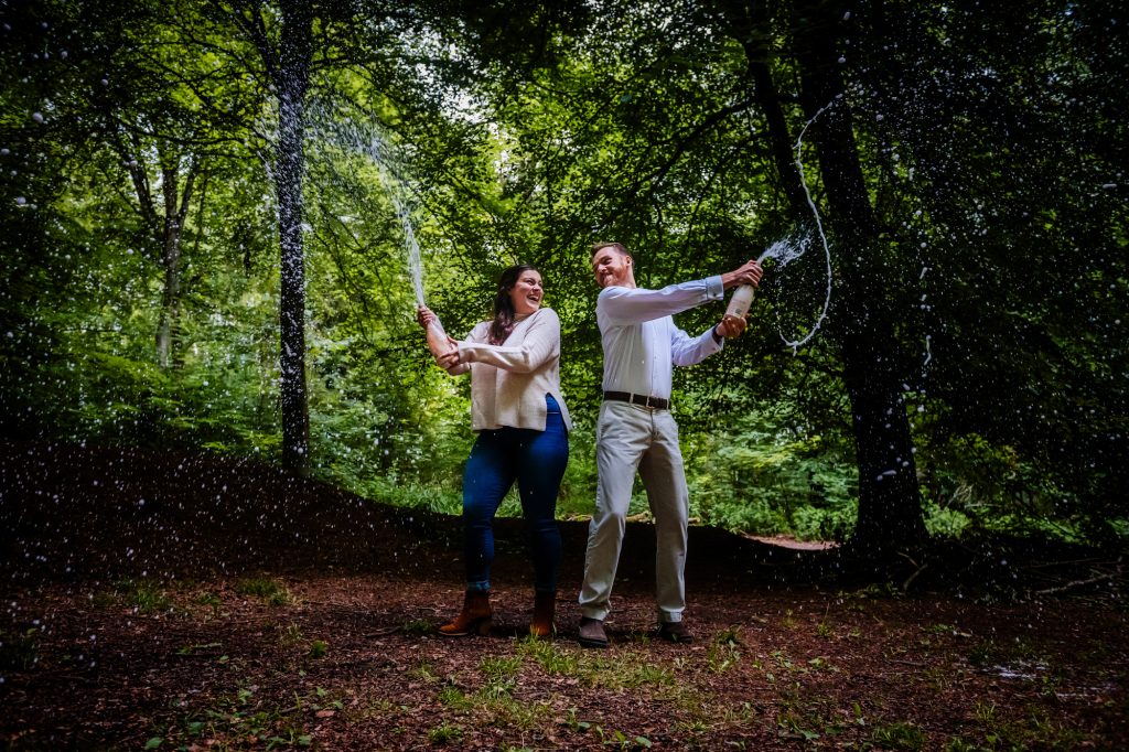 A couple playfully popping champagne in the forest during a fun pre-wedding photoshoot.
