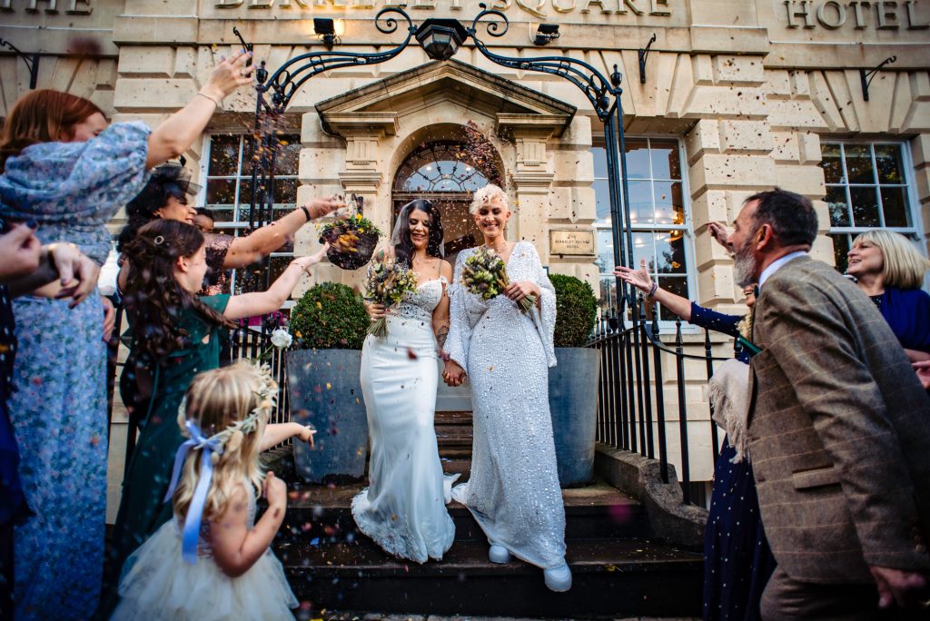 Brides walking down the steps of Berkeley Square Hotel as guests throw colourful confetti in celebration.