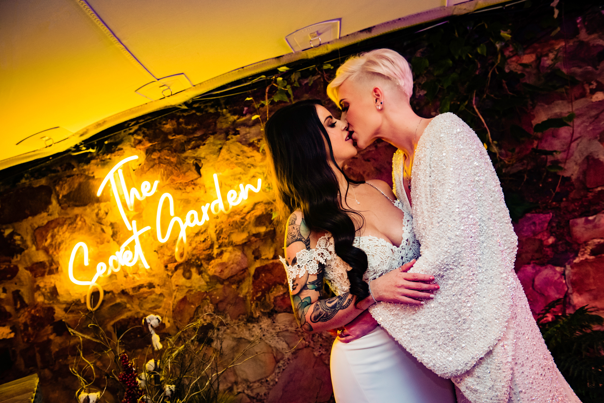 Romantic kiss between two brides under The Secret Garden neon sign at The Square Club Bristol.