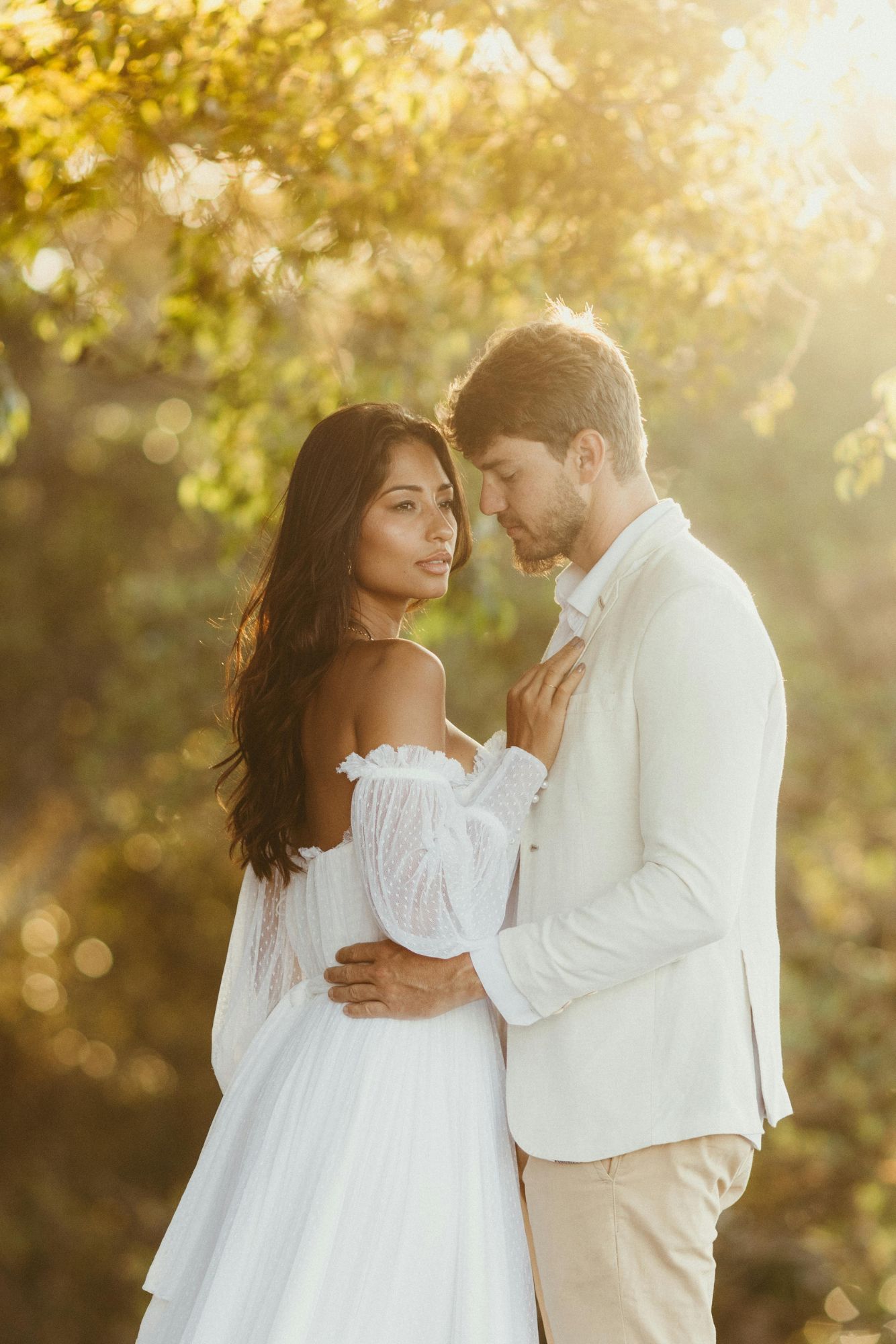 A couple in soft golden light wearing light-toned outfits during a romantic pre-wedding photoshoot outdoors.