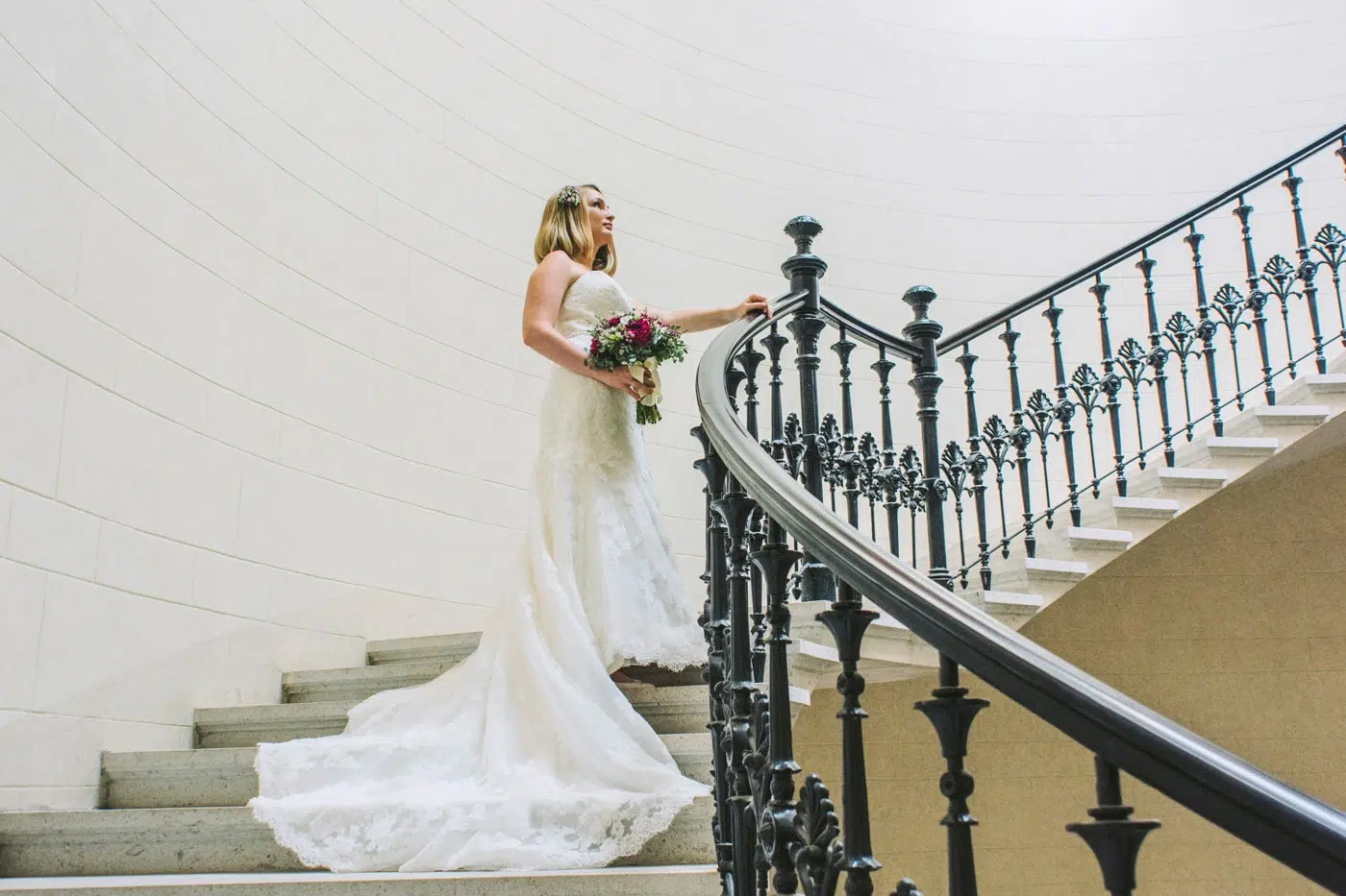 Bride walking down curved staircase in lace wedding dress holding bouquet during elegant bridal portrait.