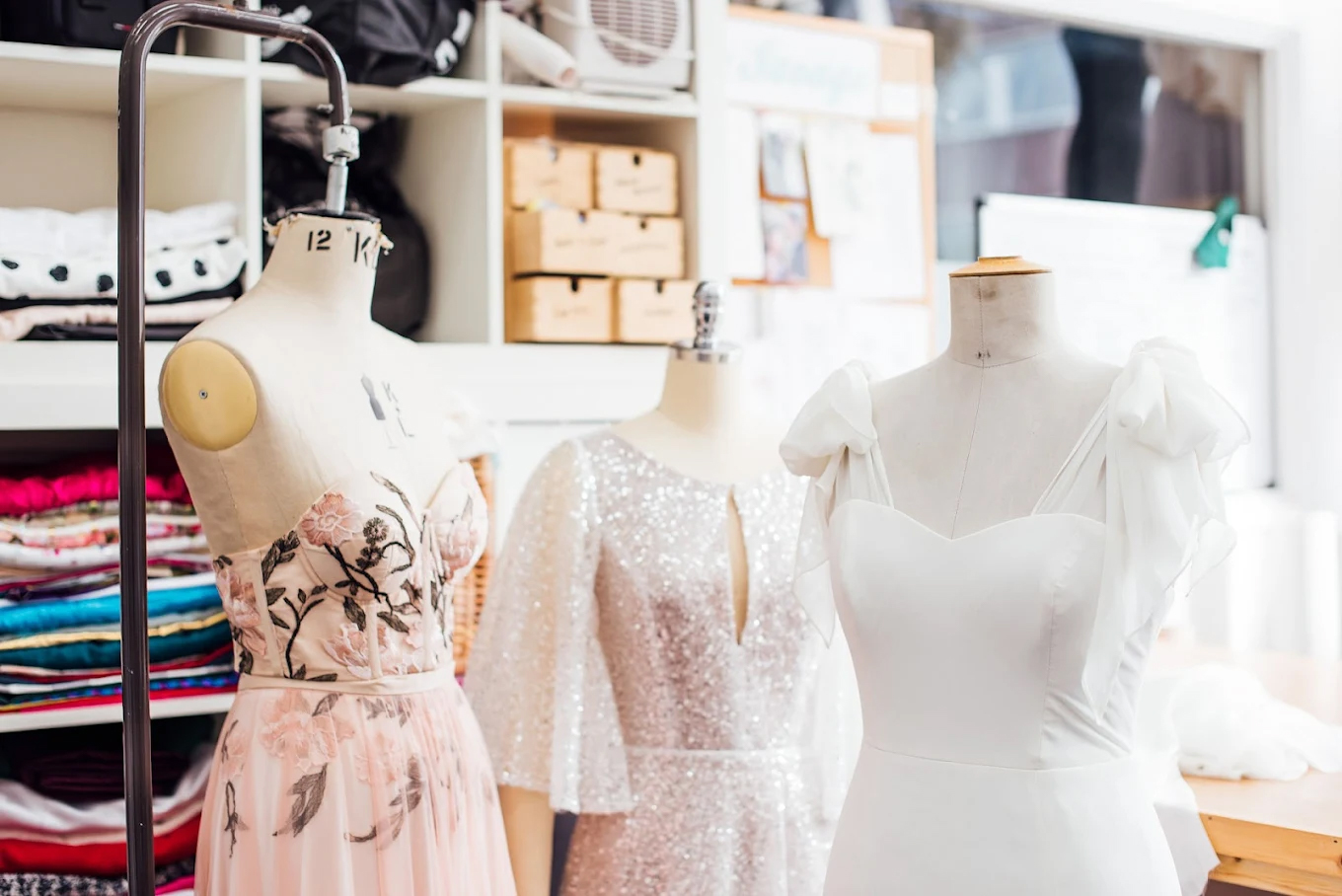 Wedding dresses displayed on mannequins inside bridal design studio with fabrics and tailoring tools.