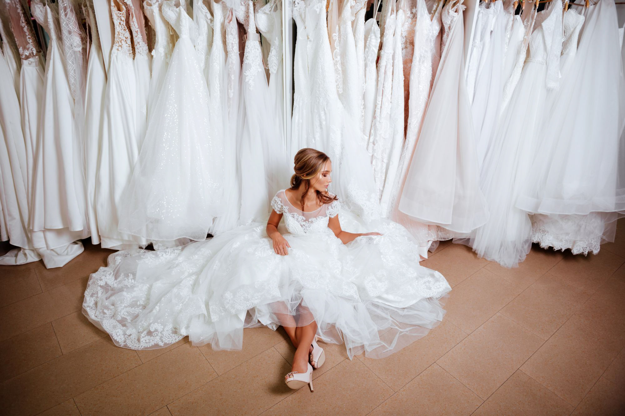 Bride sitting among wedding dresses inside bridal boutique while choosing the perfect gown.