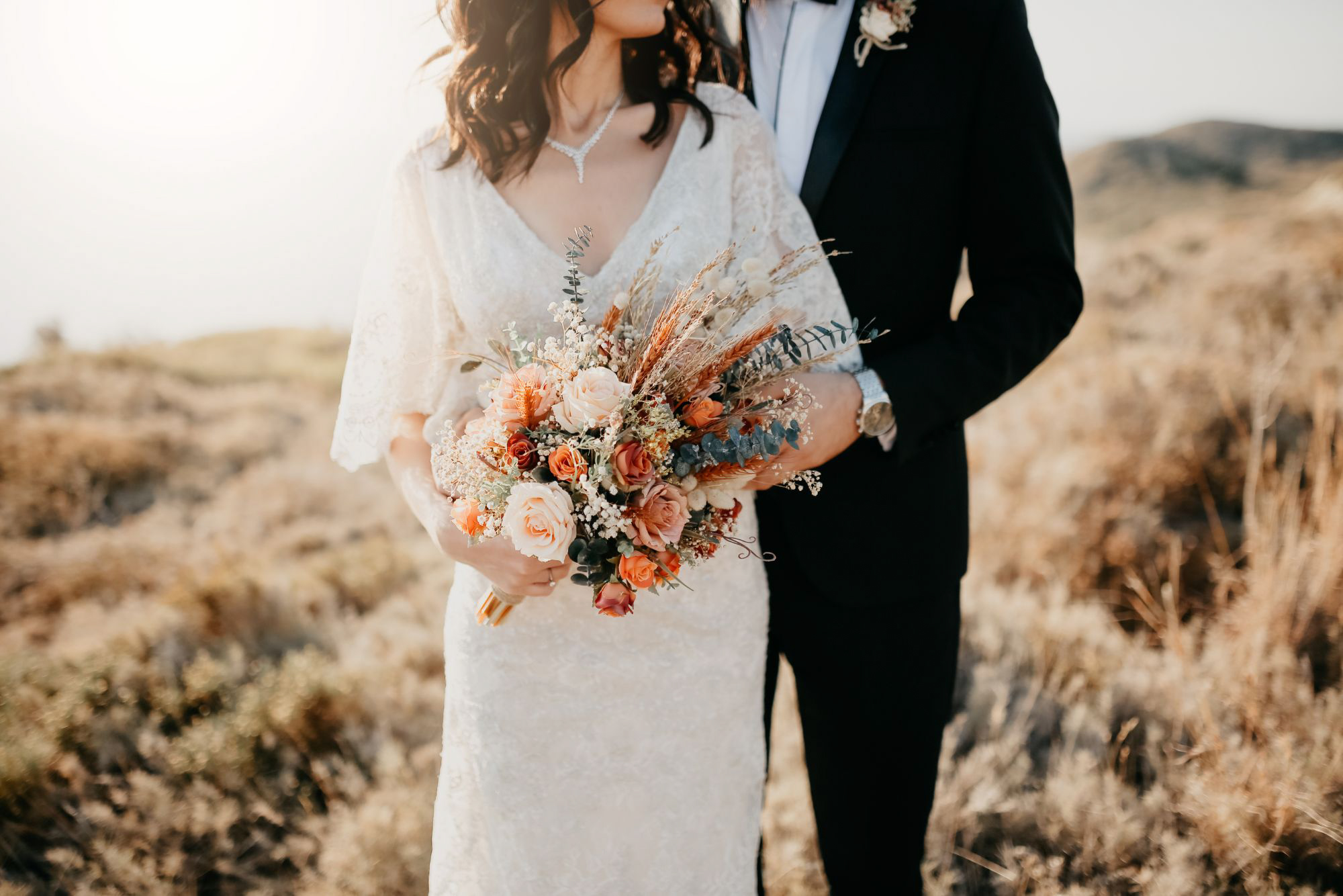 Bride in lace wedding dress holding bouquet while standing with groom during outdoor portrait.