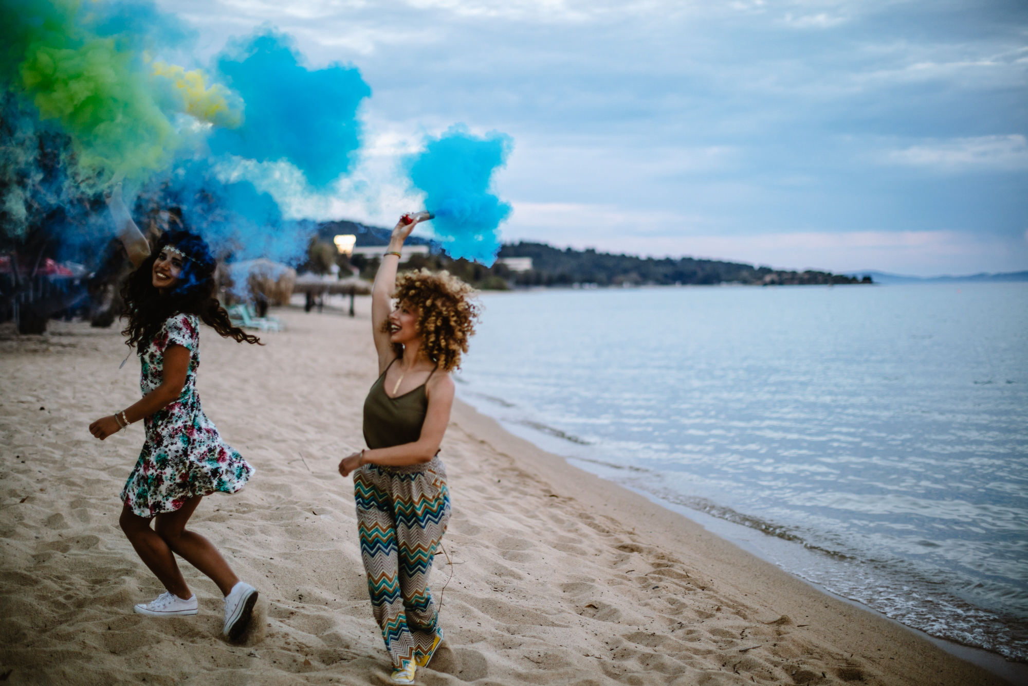 Two women run along a beach with blue and green smoke bombs under a cloudy sky.