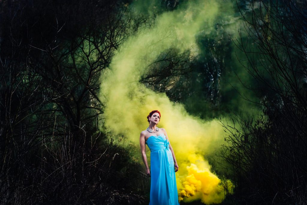 Red-haired bride in a blue dress stands among green and yellow smoke in woodland.