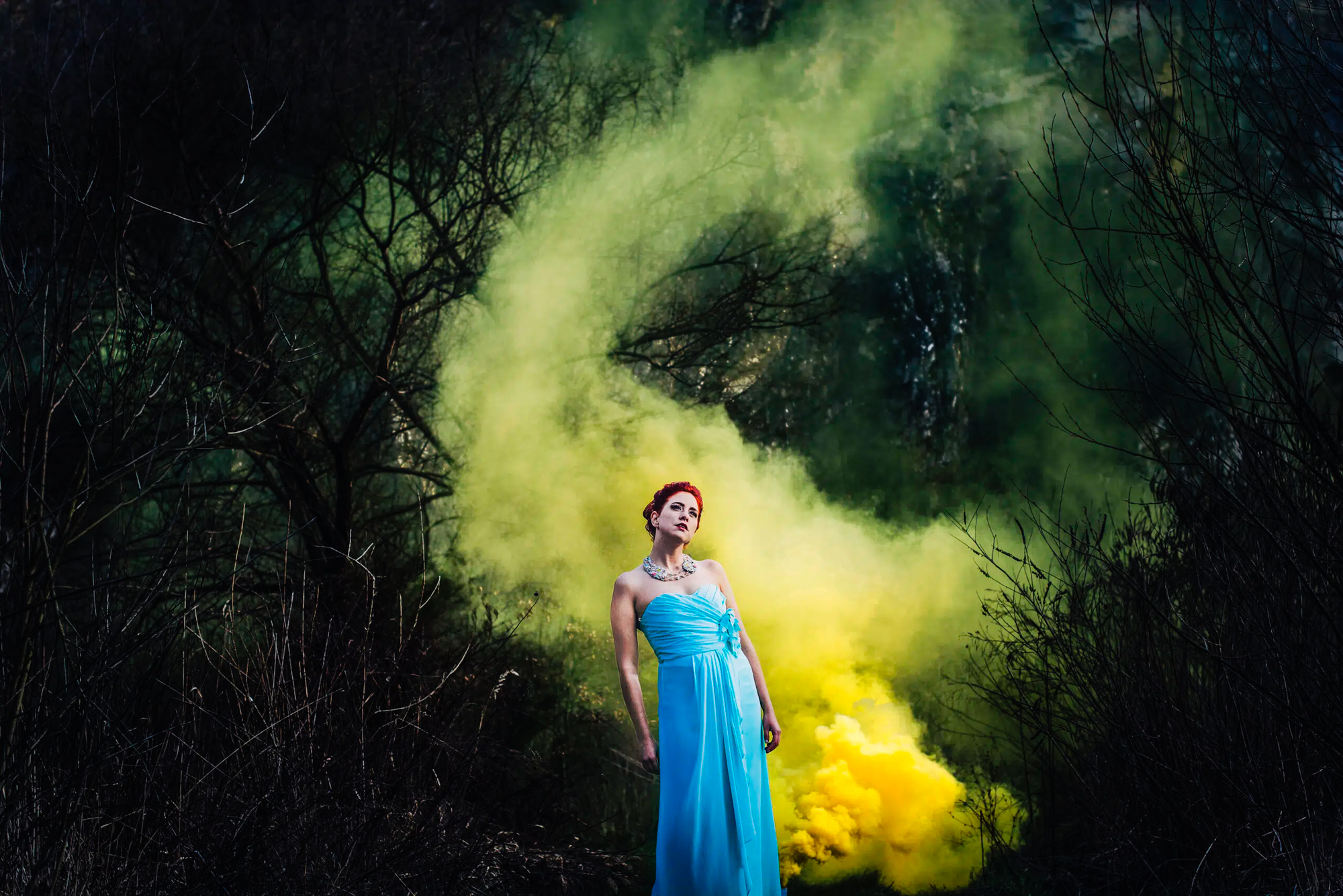 Red-haired bride in a blue dress stands among green and yellow smoke in woodland.