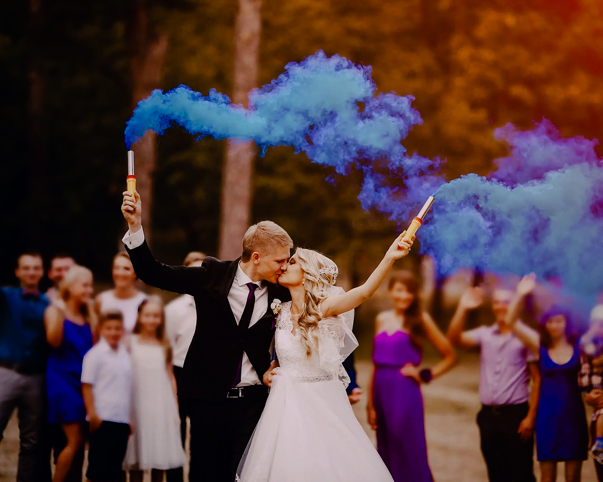 Bride and groom kiss while holding blue smoke bombs with wedding guests blurred behind them.