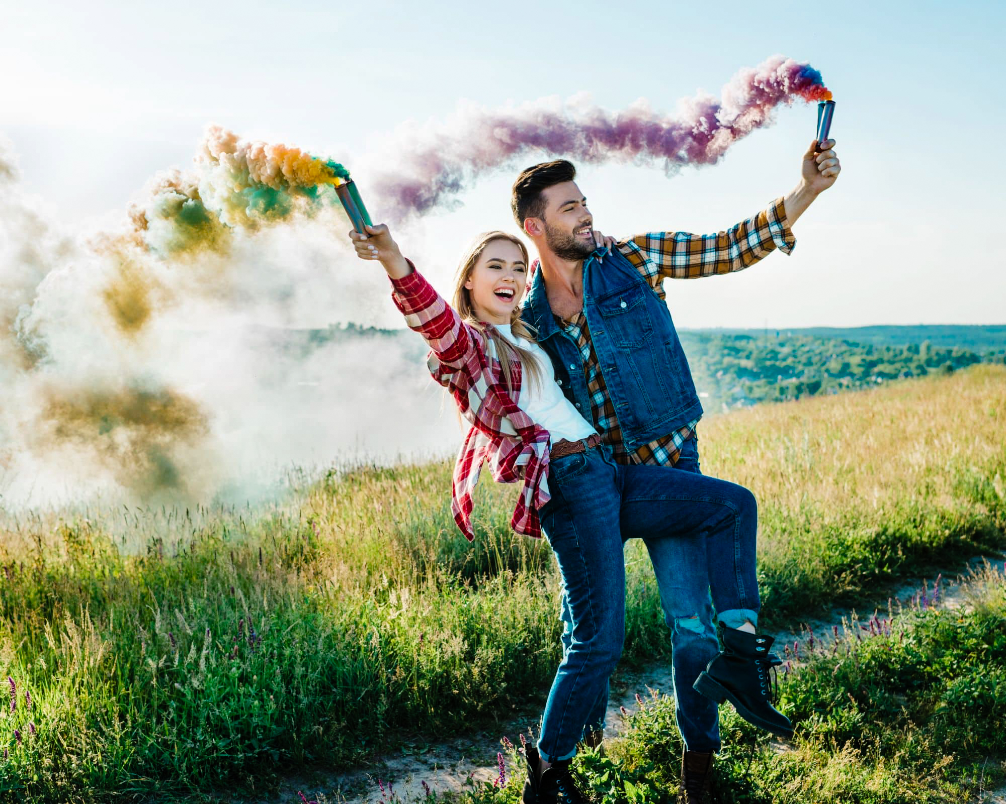 Couple laugh on a grassy hill while holding colourful smoke bombs in bright sunlight.