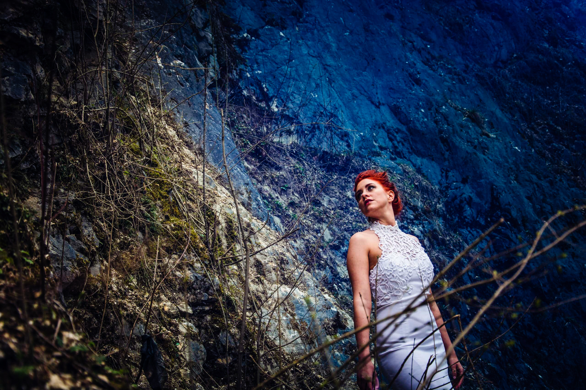 Bride in a white dress stands beside a blue rock face in dramatic moody light.