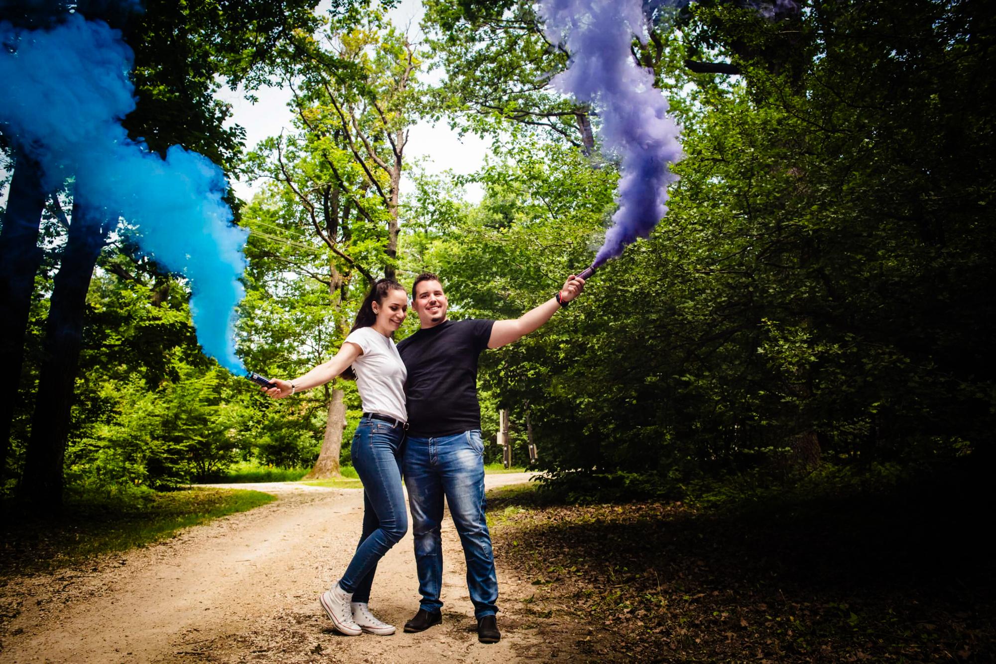 Couple pose on a woodland path holding blue and purple smoke bombs overhead.