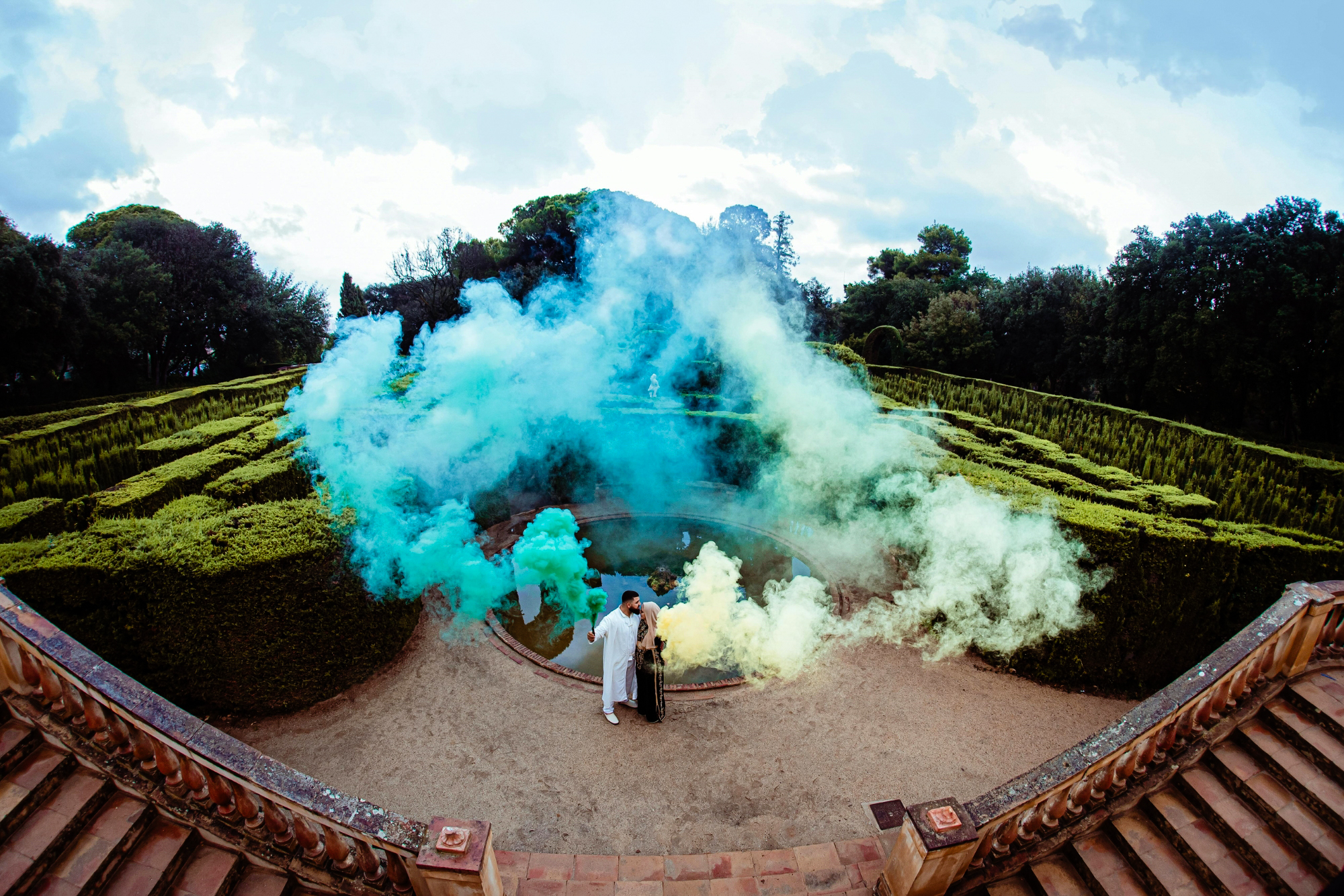Couple stands in formalwear inside a maze garden surrounded by blue, green, and yellow smoke.
