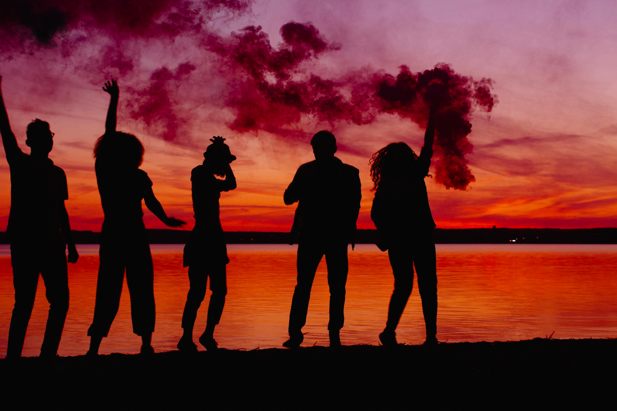 Five silhouetted figures hold red smoke bombs beside a lake at vivid sunset.