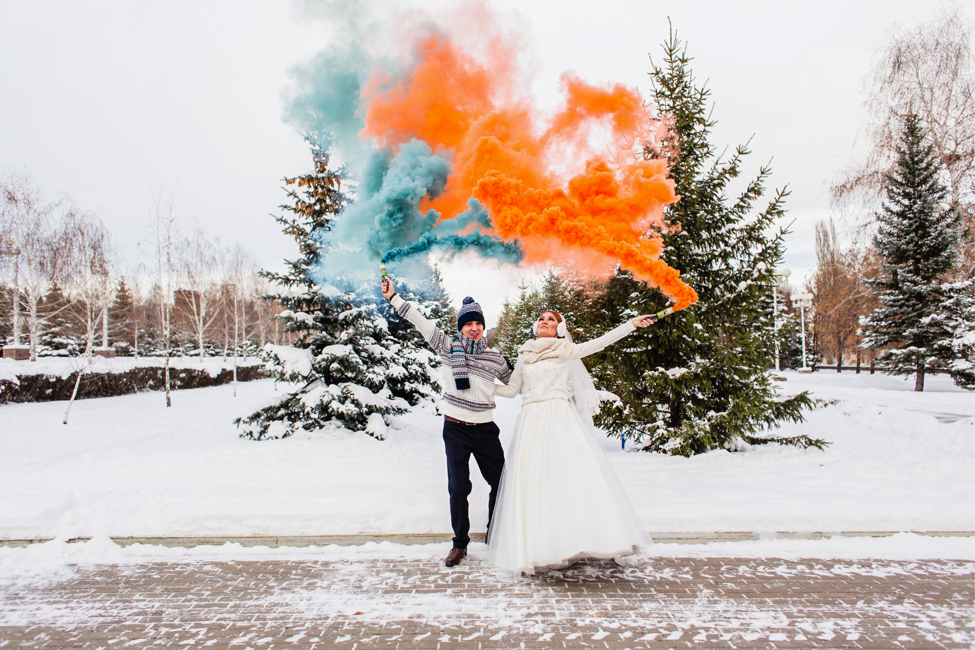 Bride and groom hold blue and orange smoke bombs in a snowy park.