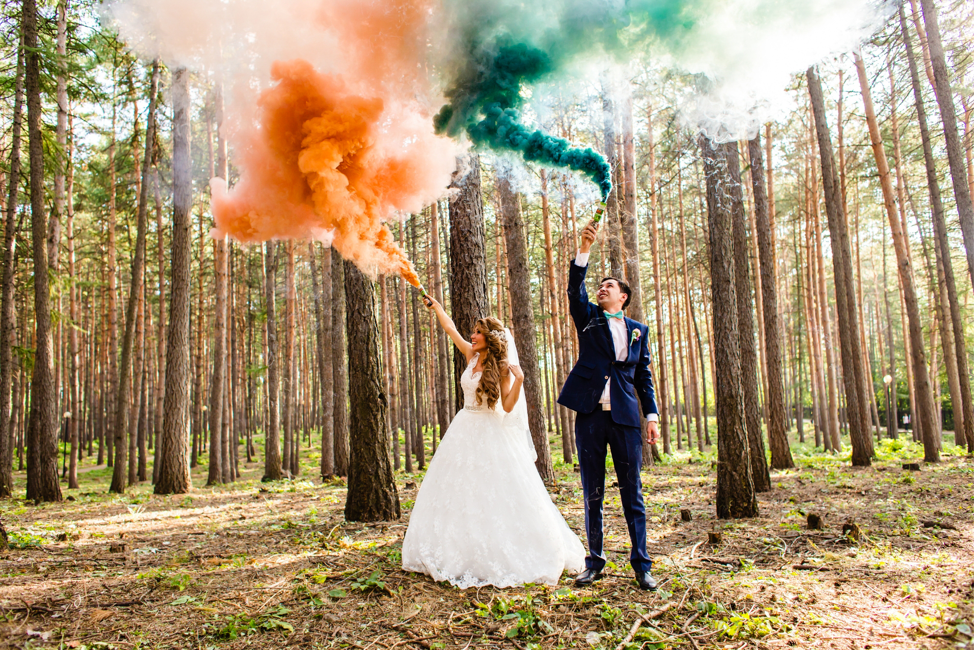 Bride and groom raise orange and green smoke bombs in a sunlit pine forest.