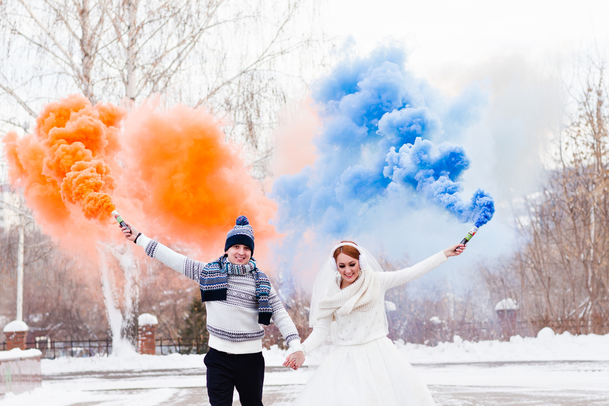 Winter couple holds hands beneath orange and blue smoke clouds in a snowy setting.