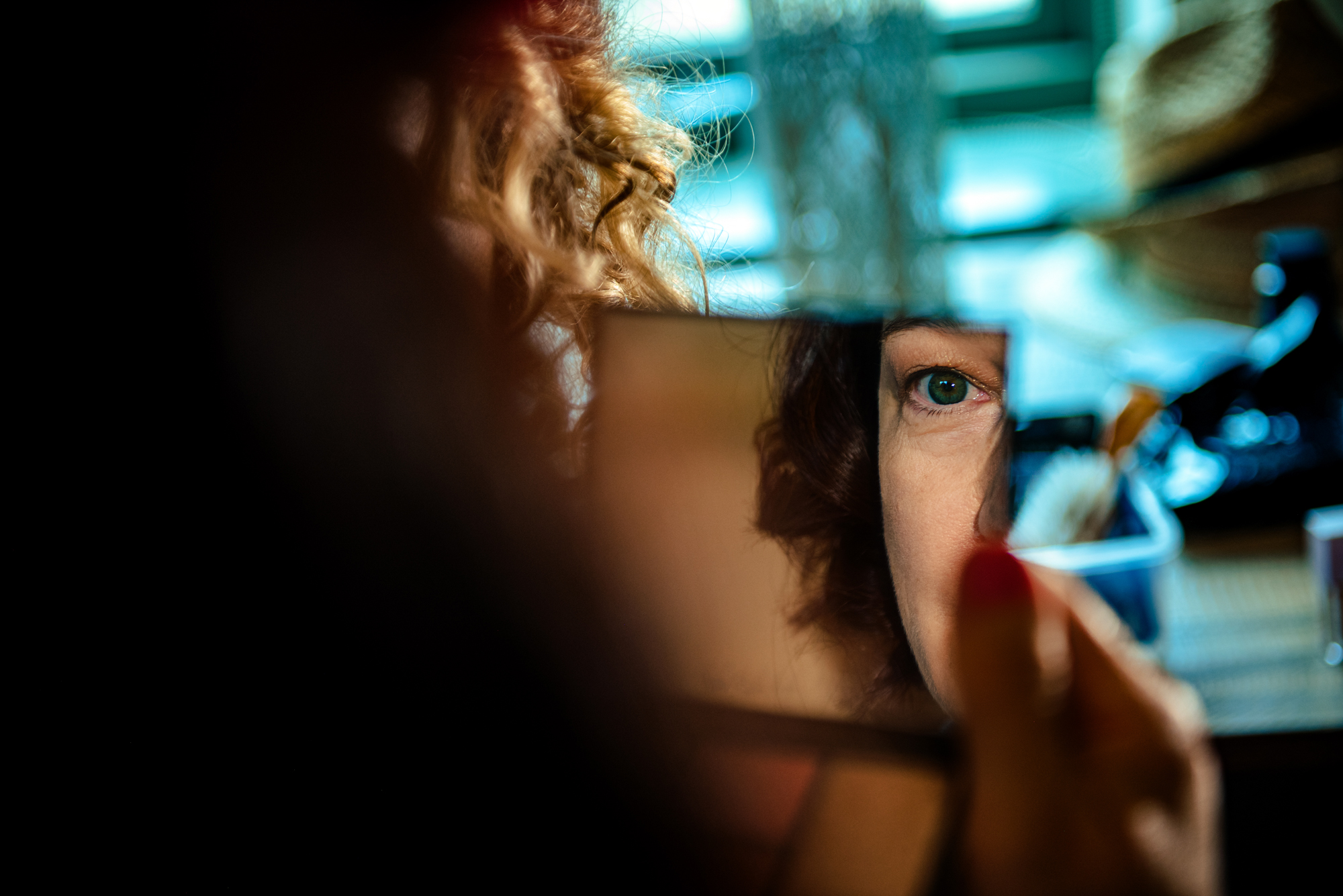A woman’s green eye appears sharply in a small handheld mirror while she perfects her makeup.