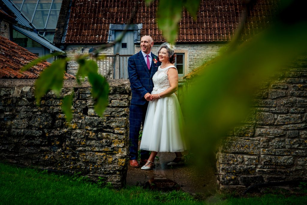 The couple stand close beside the old stone wall, partially hidden by soft green leaves in the foreground.