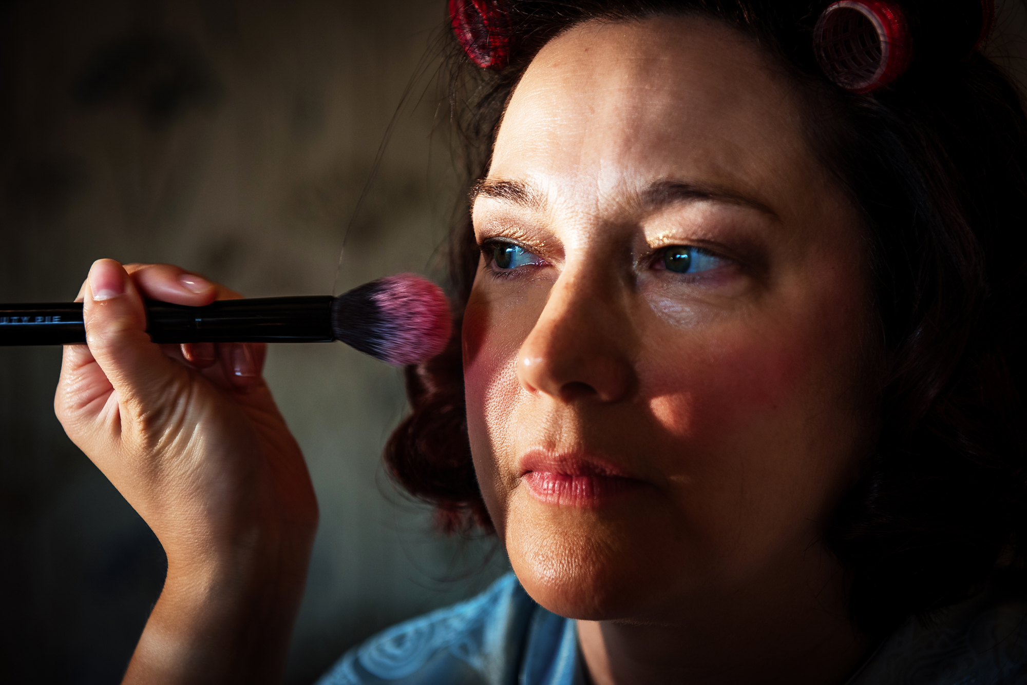 Soft morning light highlights her face as blusher is gently applied during bridal preparations at Folly Farm.