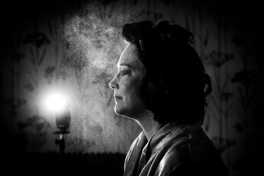 A mist of setting spray fills the air as she sits calmly, lit by a bright backlight.