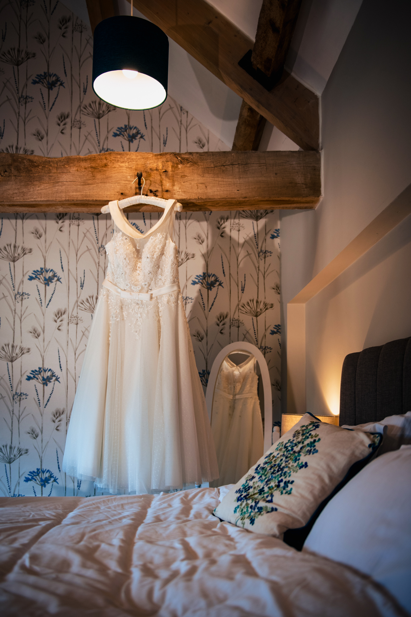 The wedding dress hangs from a wooden beam, reflected softly in the mirror beneath it.