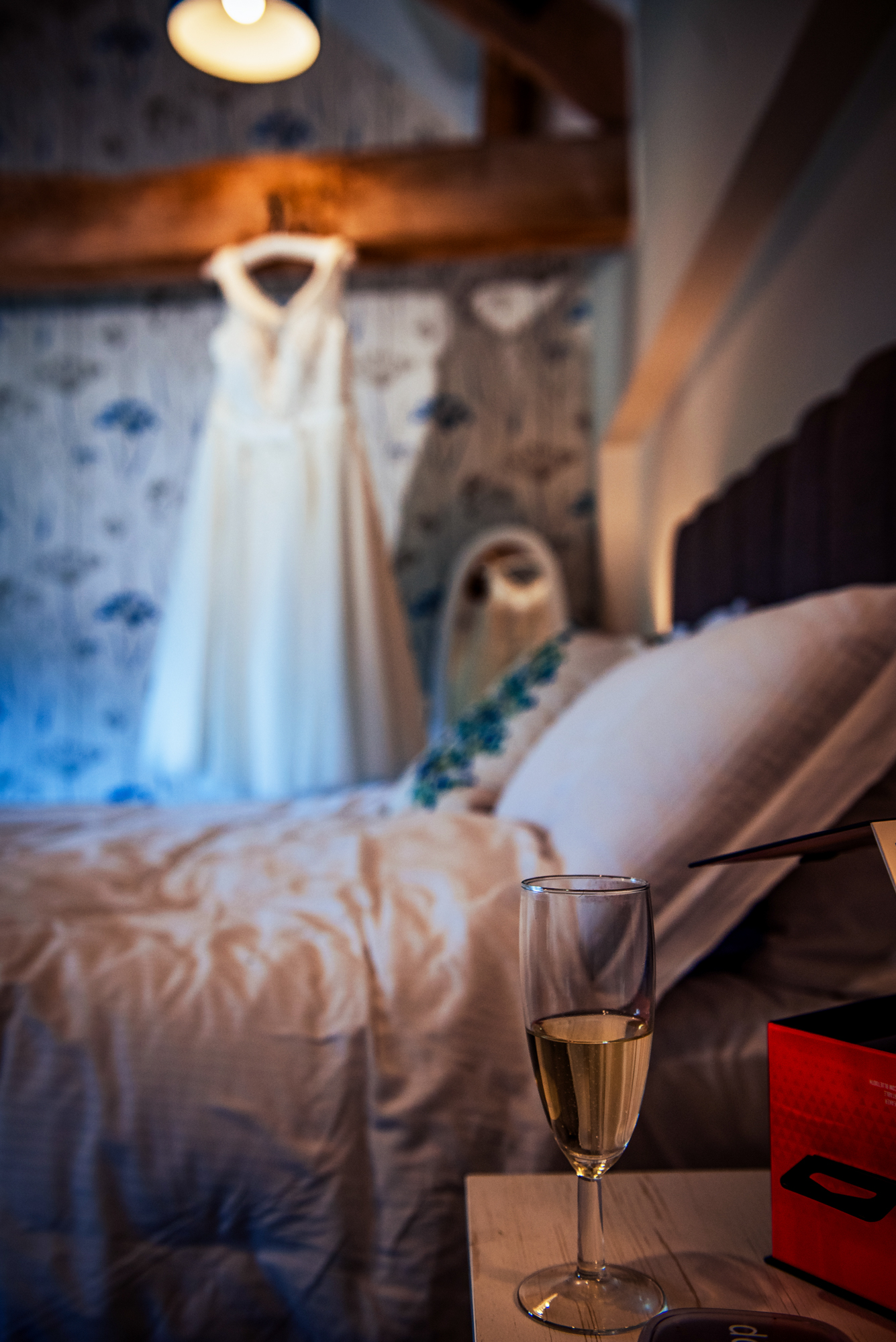 A glass of champagne rests on the bedside table while the wedding dress hangs in the background.