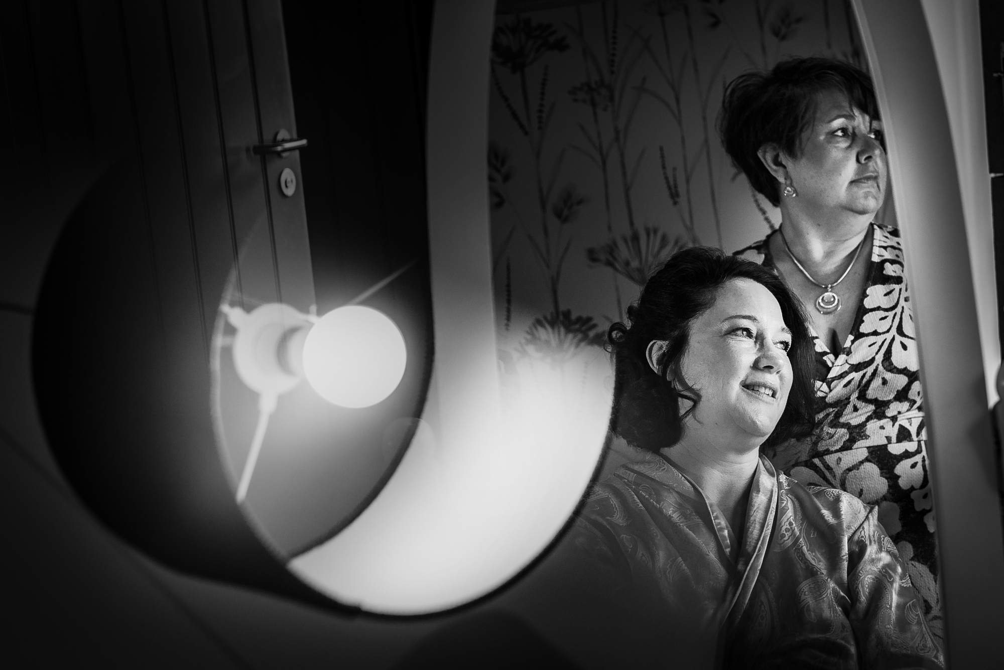 Bride smiles in the mirror beside her hairstylist, surrounded by soft light and patterned wallpaper.