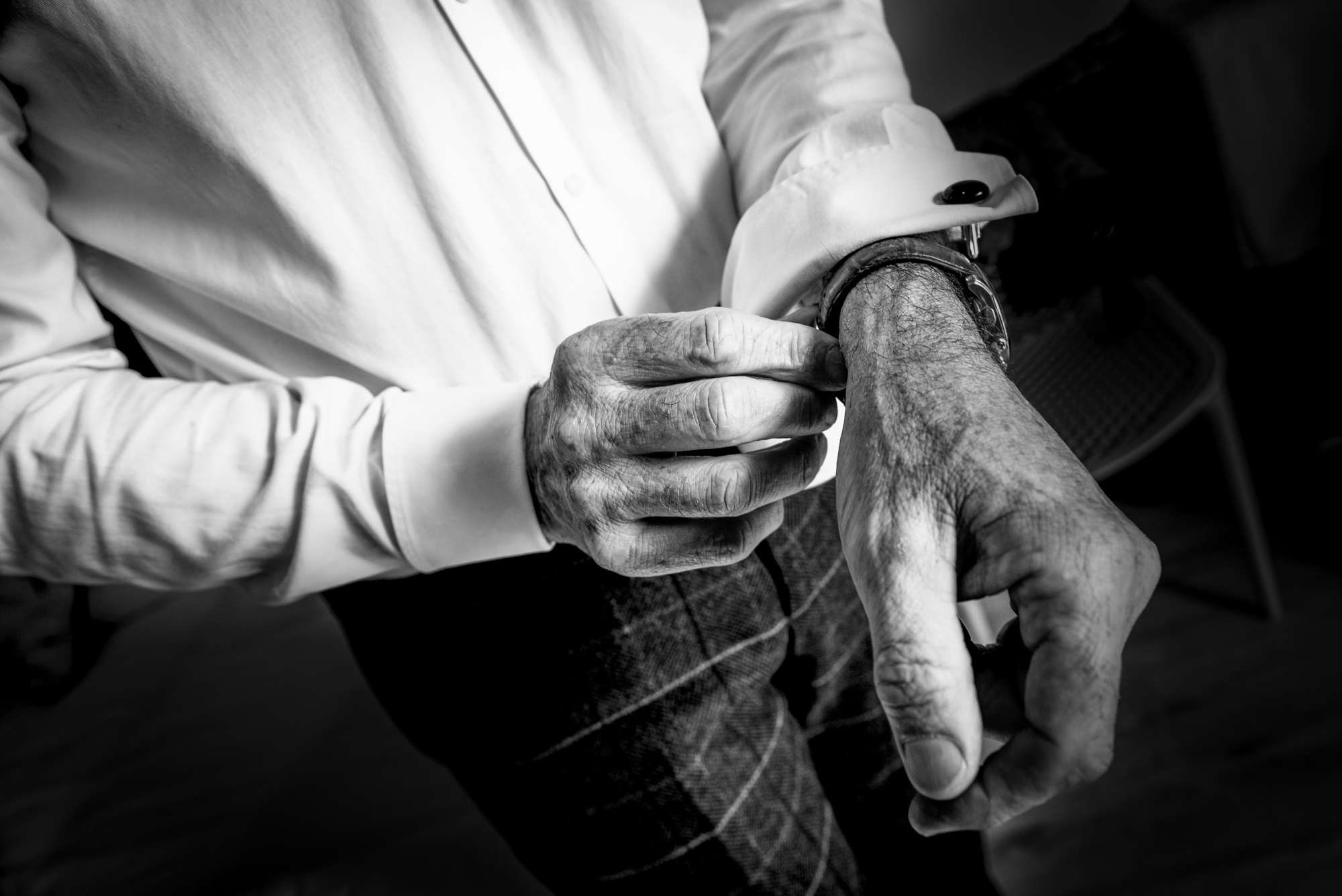 Hands adjust a cufflink and watch during groom preparations, showing textured detail in monochrome.