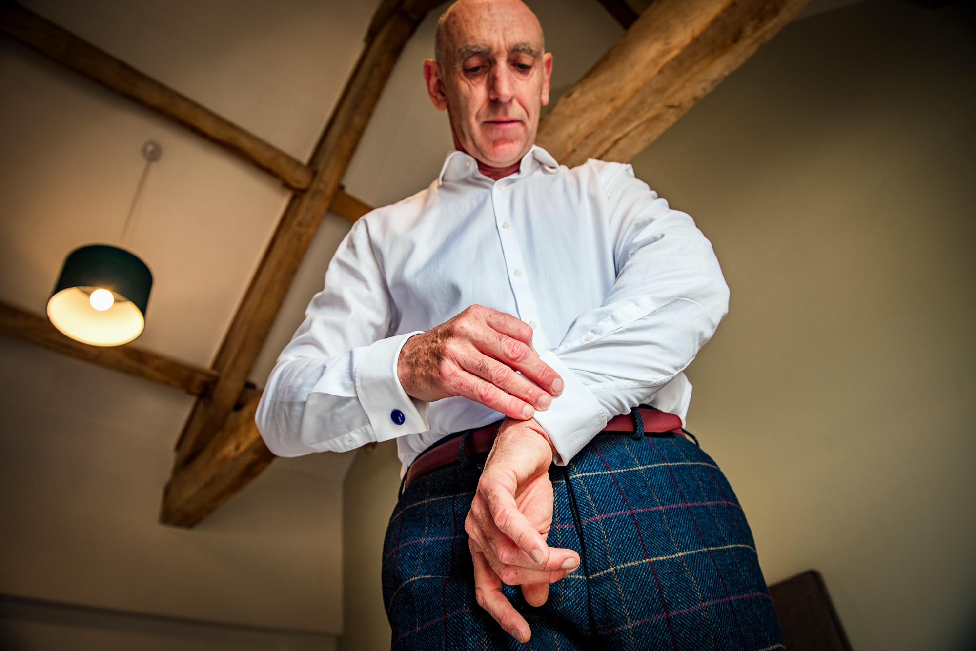 The groom fastens his cufflinks beneath the wooden beams, wearing checked trousers for the Scottish-inspired wedding.