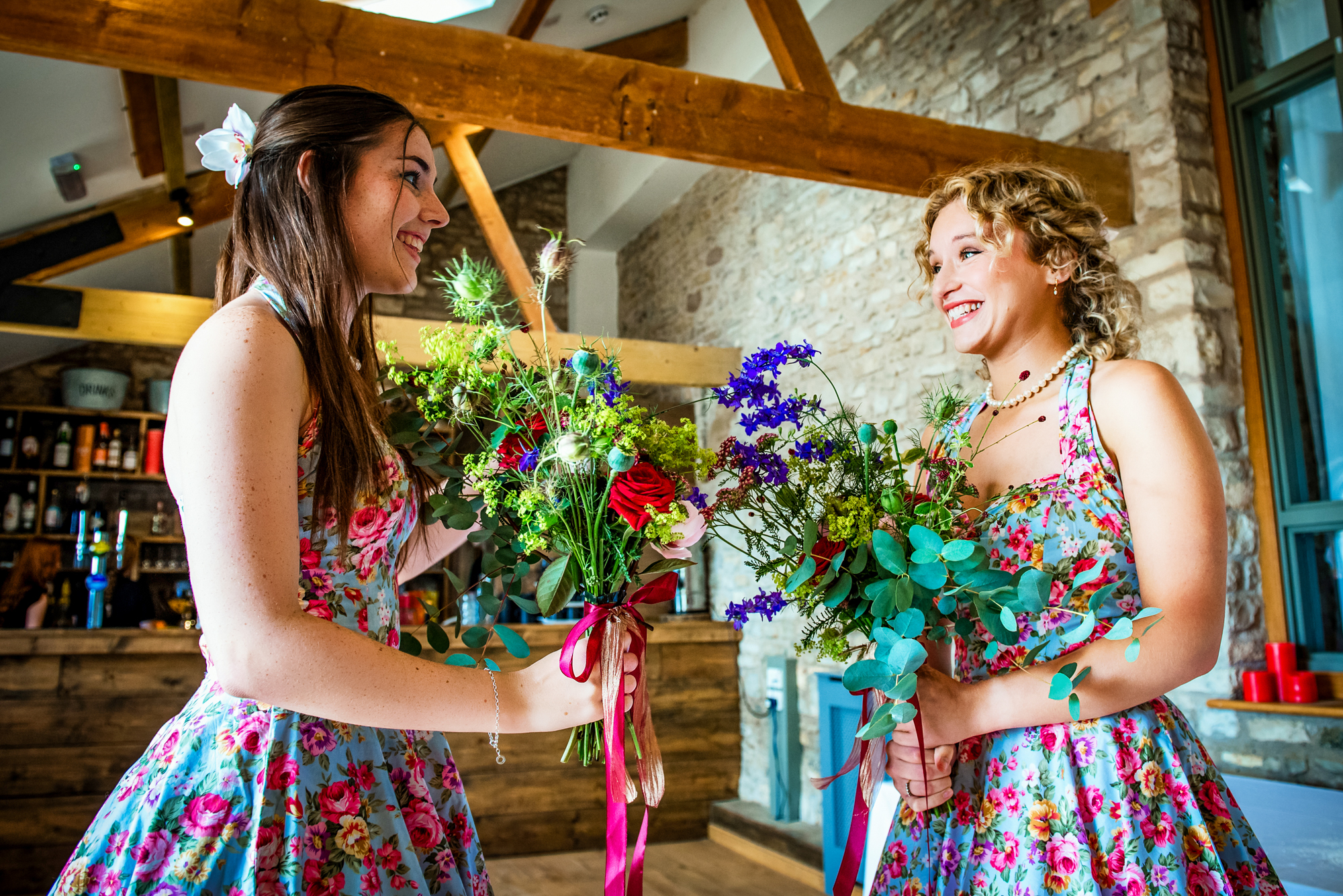 Two bridesmaids smile and compare their colourful bouquets inside Folly Farm’s rustic bar area.