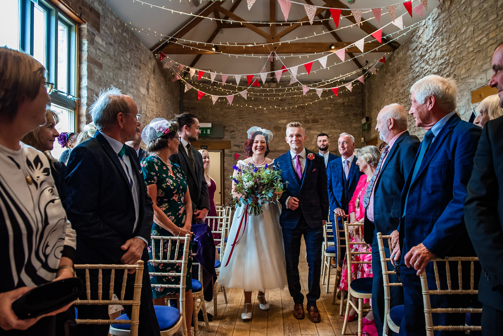 The bride walks down the aisle smiling, surrounded by guests and bunting decorations.