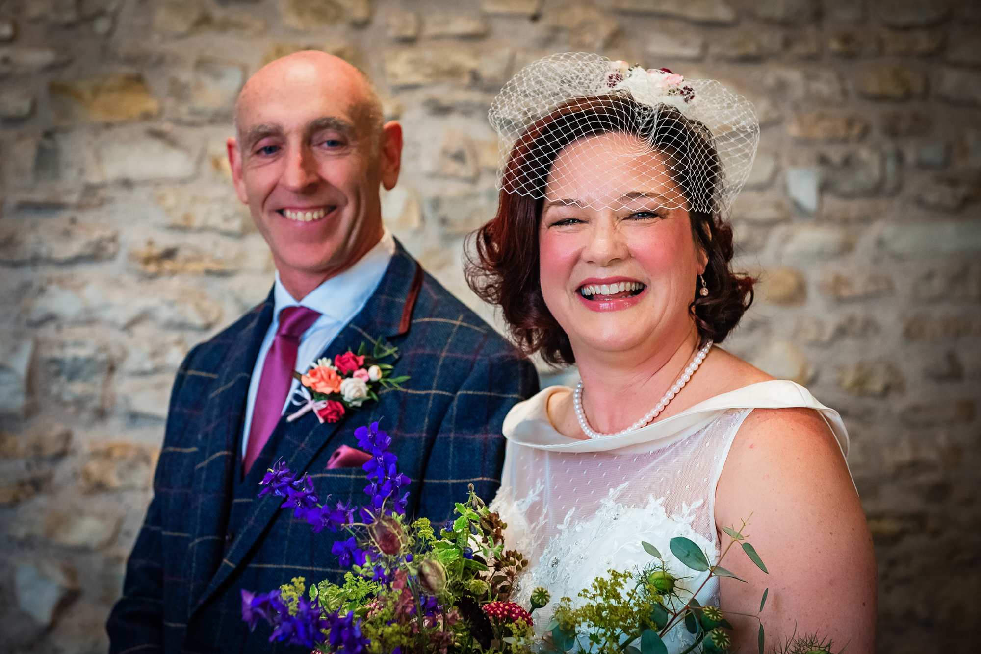 The couple stands close together, smiling brightly with wildflowers and stone textures behind them.