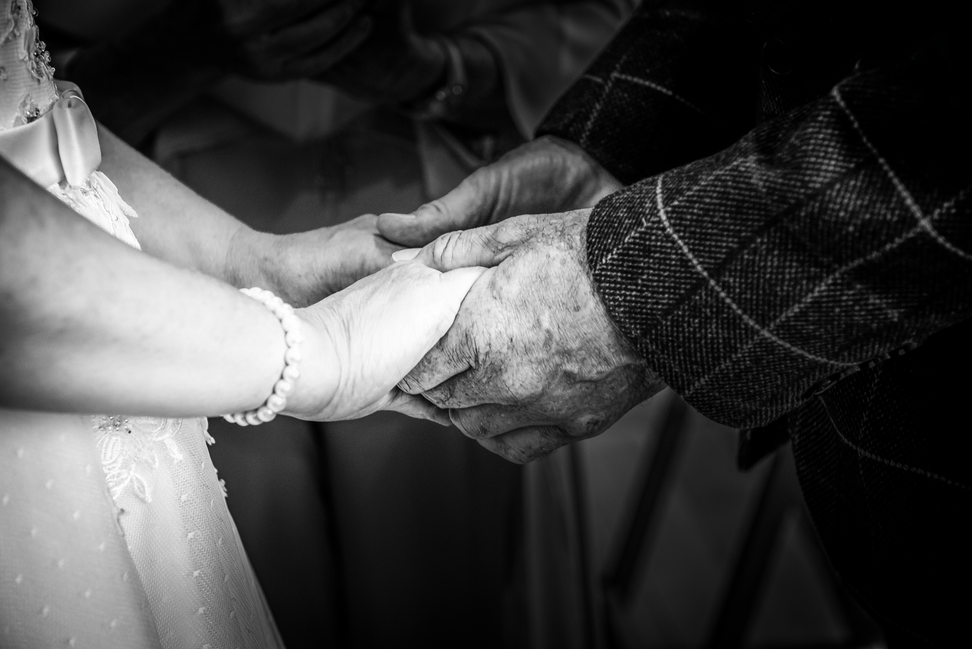 Their hands rest together softly during the vows, showing lace, pearls, and a checked jacket sleeve.