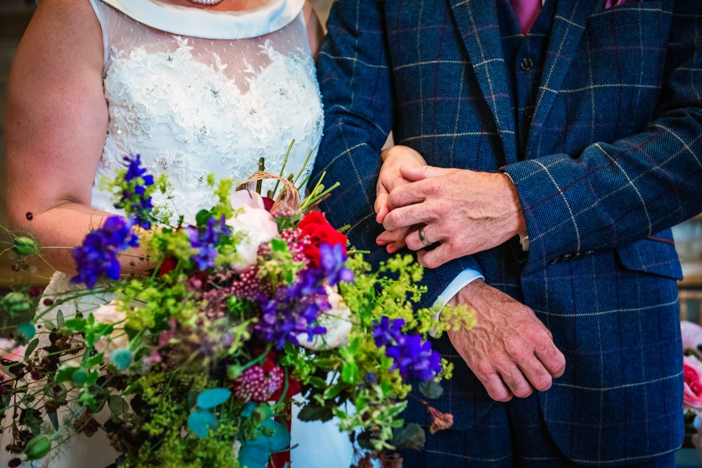 Their hands rest together beside a colourful bouquet of wildflowers against the bride’s lace dress.
