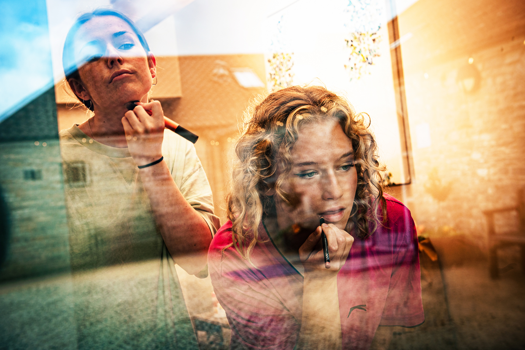 Two women apply makeup by the window, their reflections blending with Folly Farm’s stone courtyard outside.