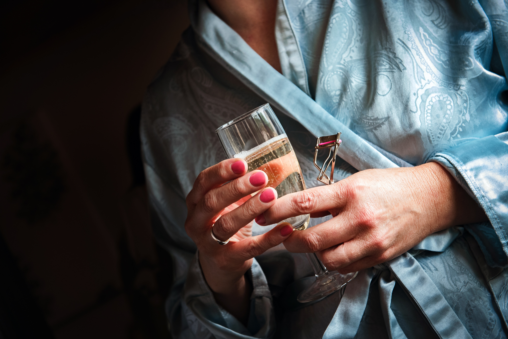 A hand holds a glass of prosecco and an eyelash curler, the pale blue robe catching soft morning light.