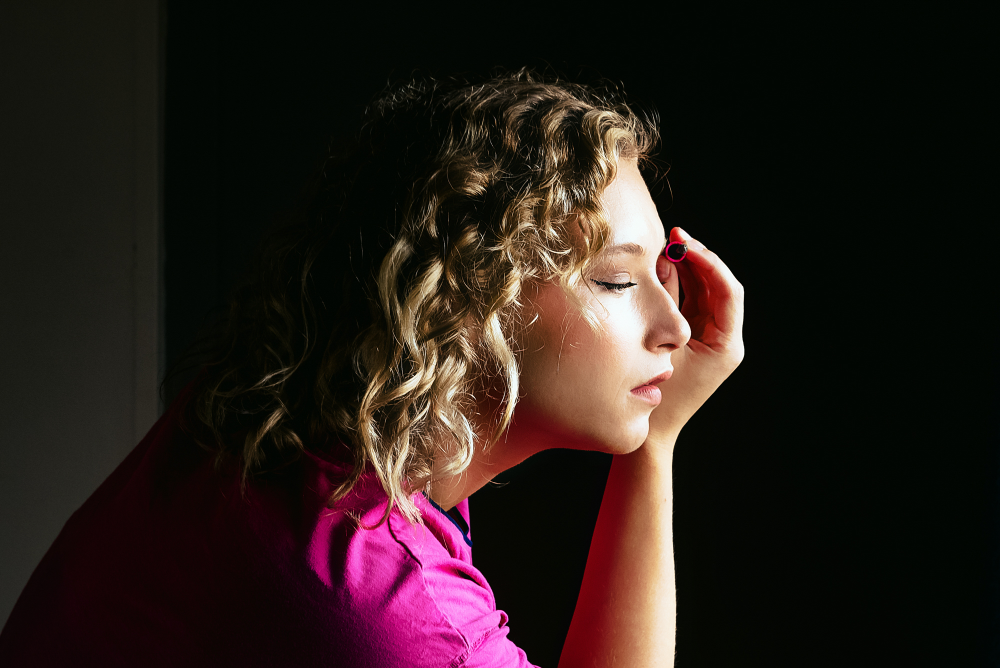 A bridesmaid pauses mid-makeup, eyes closed in calm concentration as soft side light highlights her curls.