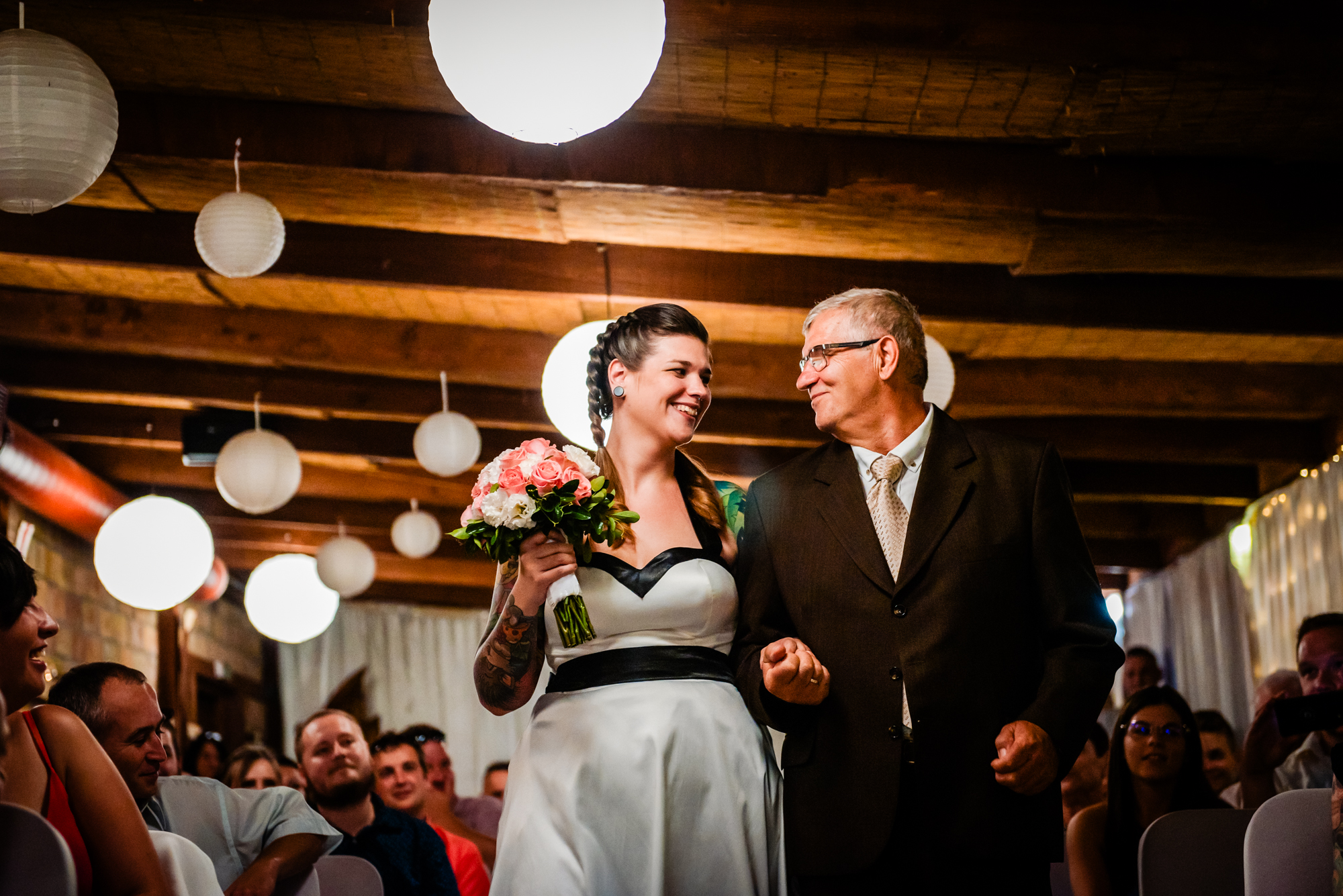 Bride and father smiling at each other while walking through warmly lit wedding ceremony aisle.