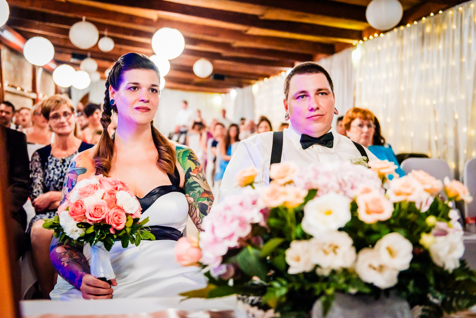 Tattooed bride and groom seated together during emotional indoor wedding ceremony with floral foreground.