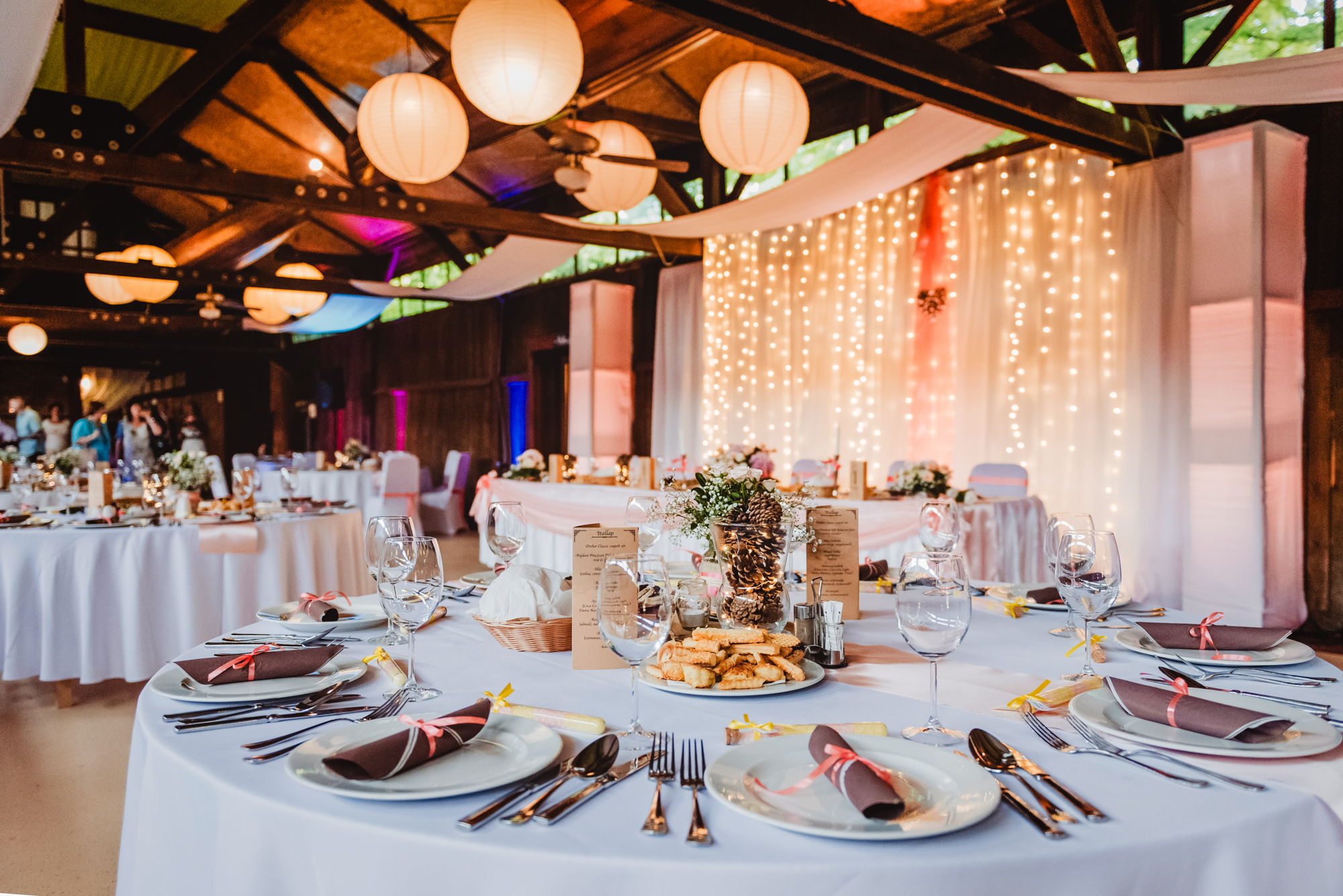 Rustic wedding reception setup with round tables, lantern lights, and fairy light backdrop at Csónakház Budapest.