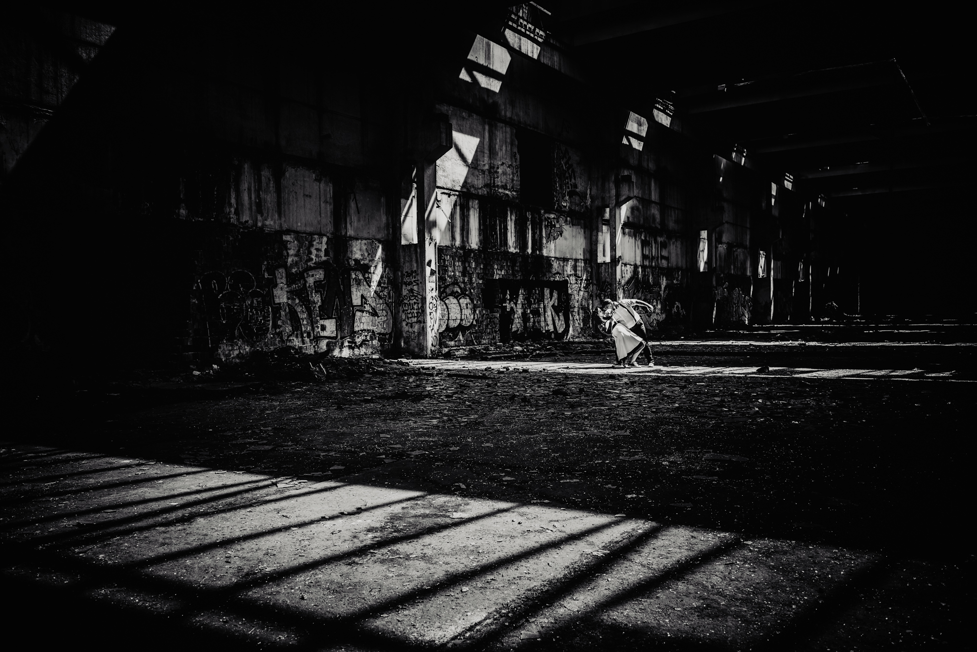 Black and white portrait of alternative newlyweds standing in dramatic light inside an abandoned industrial warehouse.