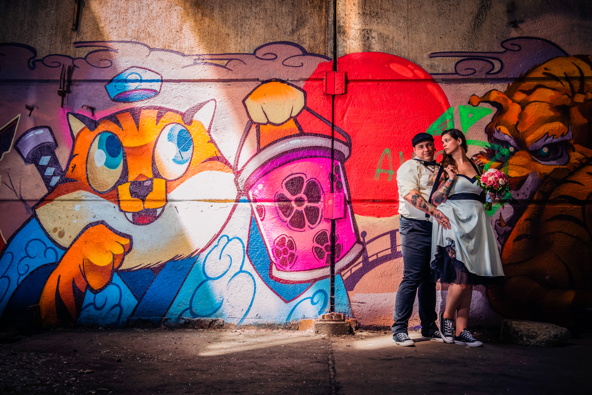 Alternative bride and groom posing beside graffiti wall with dramatic sunlight inside abandoned factory.