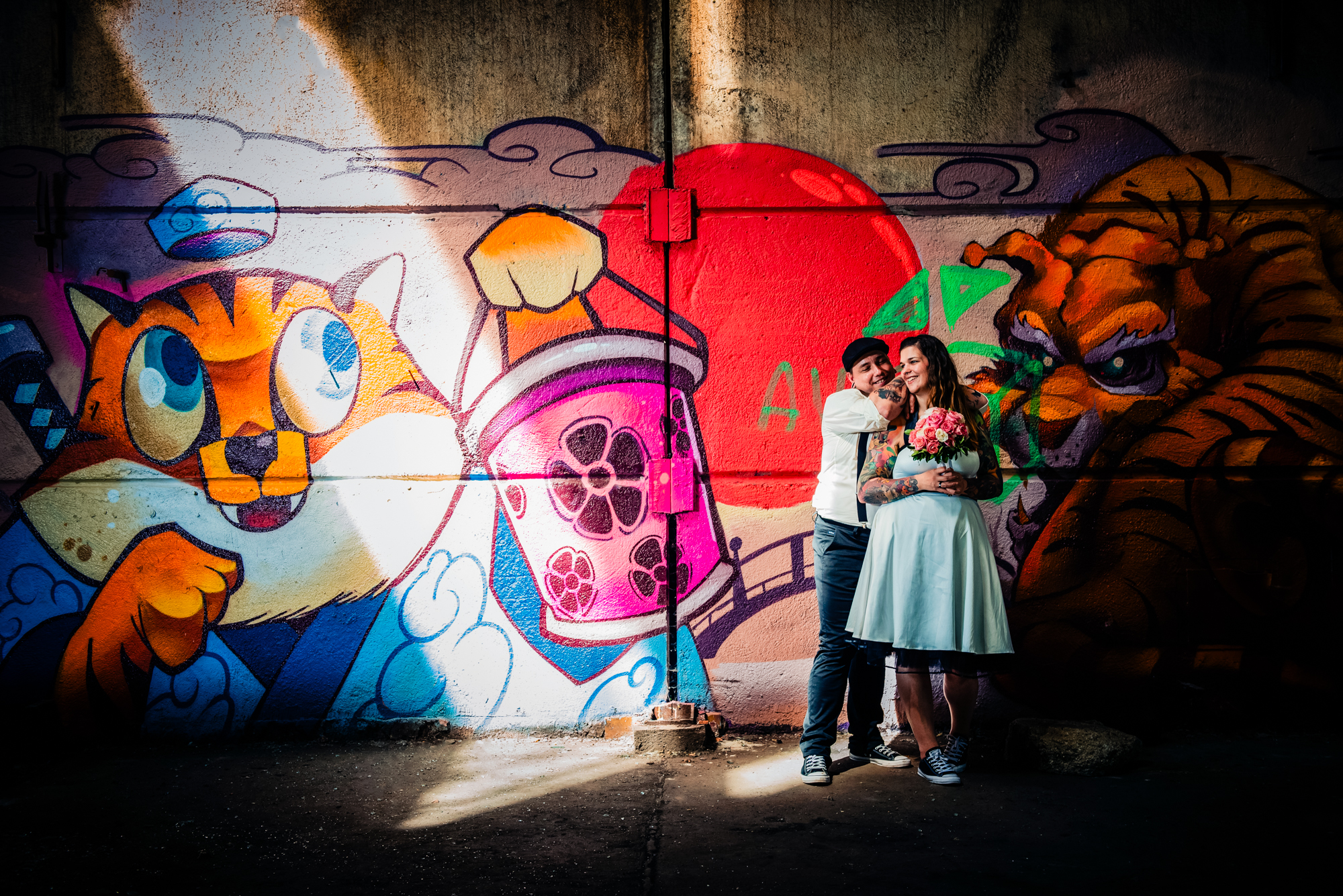 Tattooed couple smiling together in front of colourful graffiti during alternative Budapest wedding session.