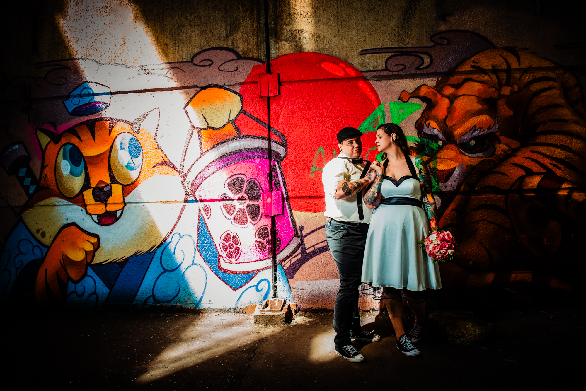 Alternative newlyweds holding hands beneath dramatic lighting and graffiti artwork in abandoned warehouse.