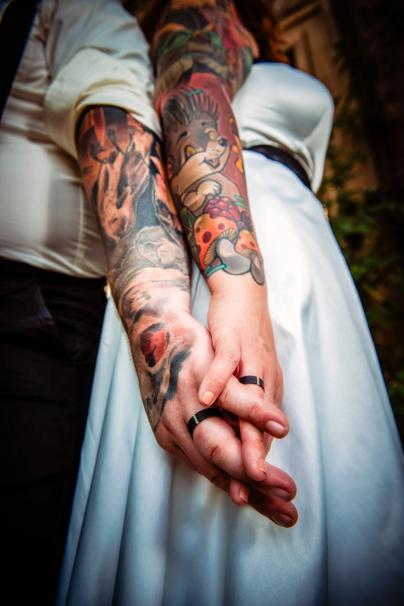 Close-up of tattooed newlyweds holding hands with black wedding bands during alternative wedding portraits.