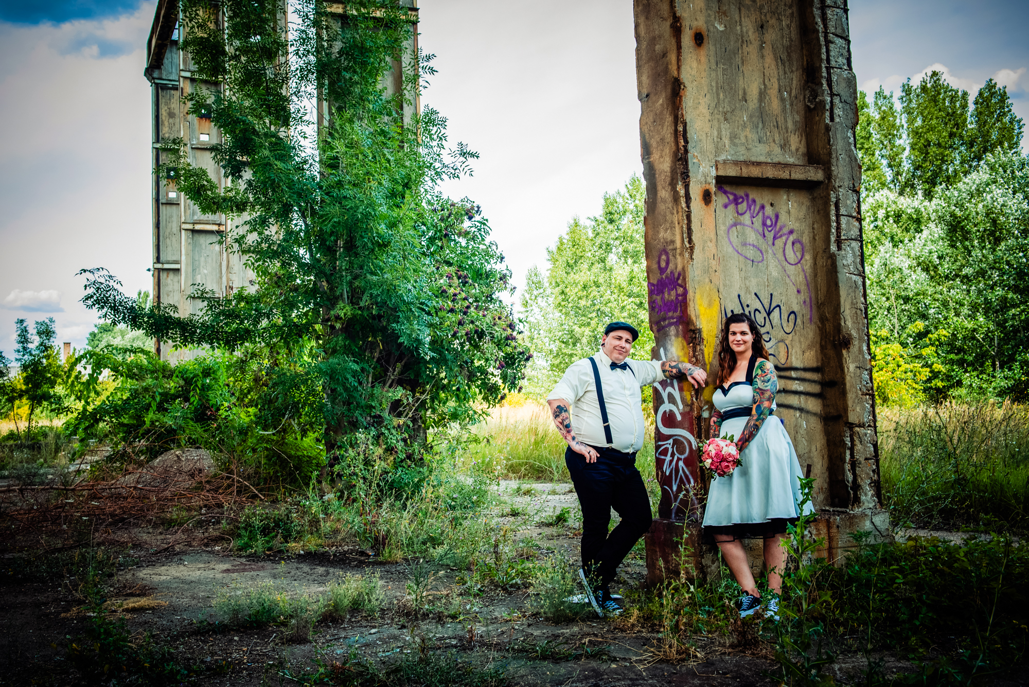 Tattooed couple posing beside graffiti-covered concrete pillar in overgrown industrial landscape near Budapest.