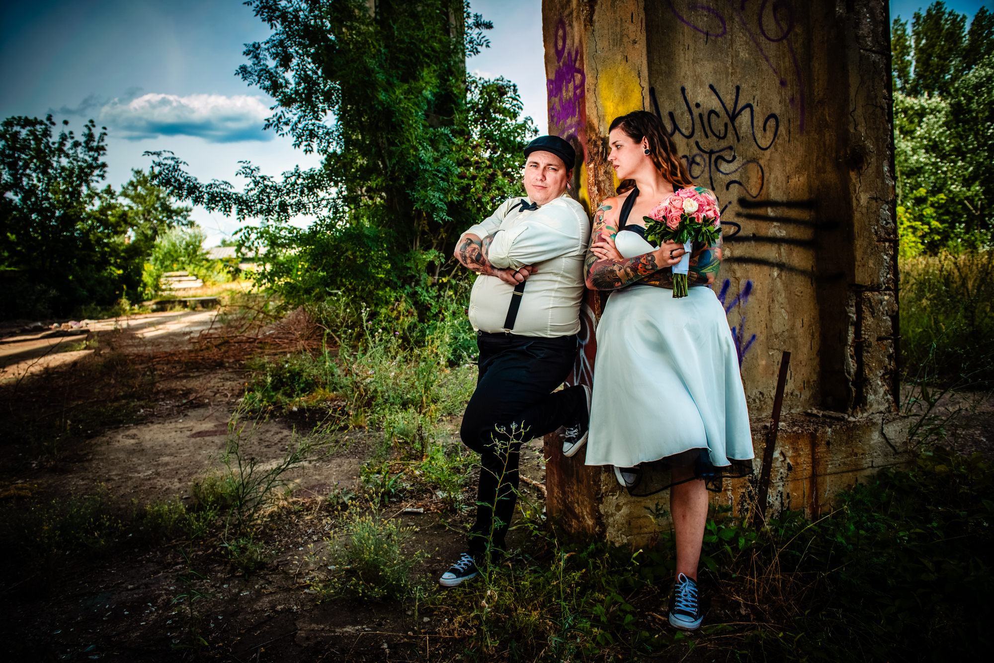 Alternative bride and groom leaning against industrial pillar surrounded by greenery and graffiti in Budapest.