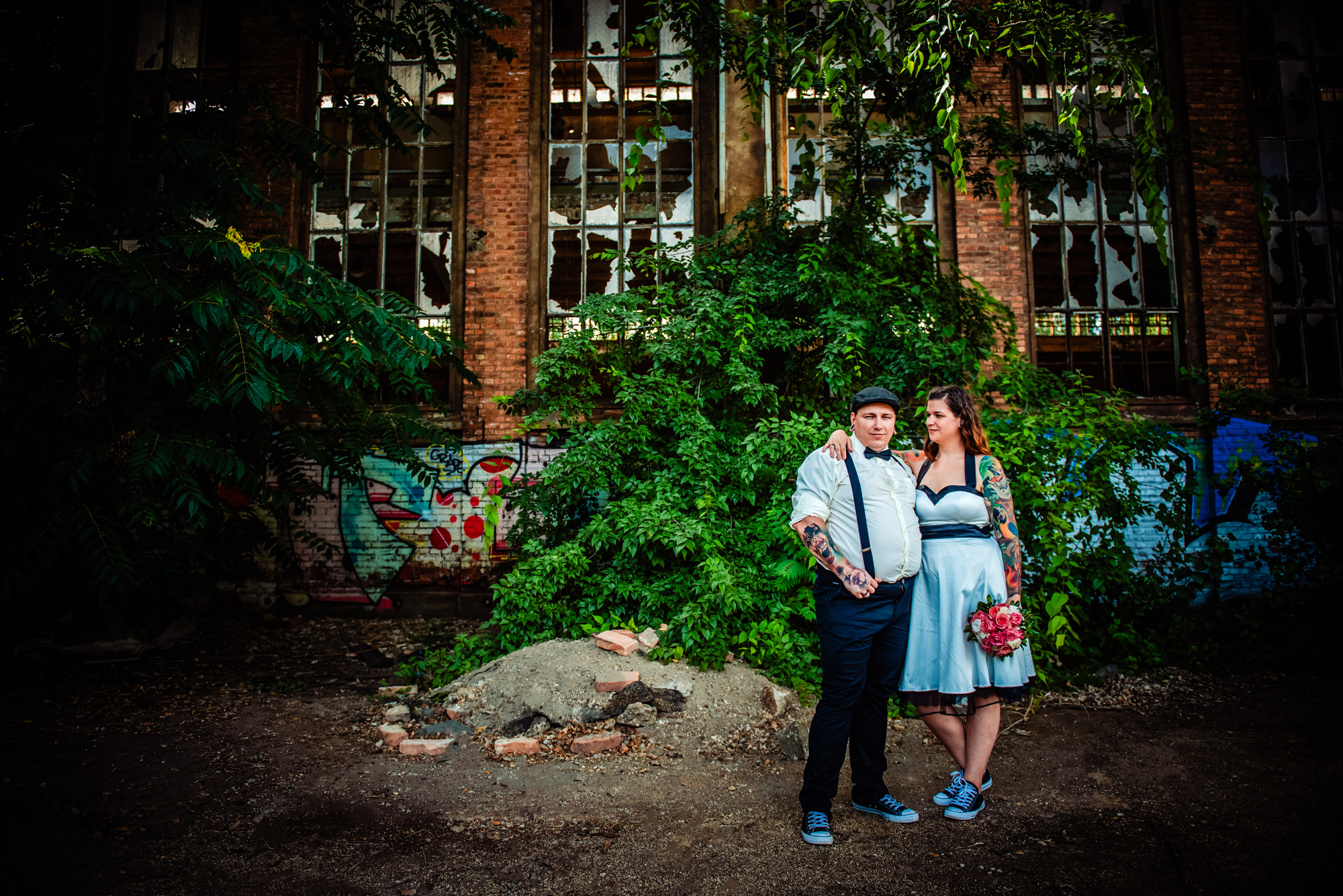 Tattooed couple standing together inside abandoned factory overtaken by plants and broken windows.