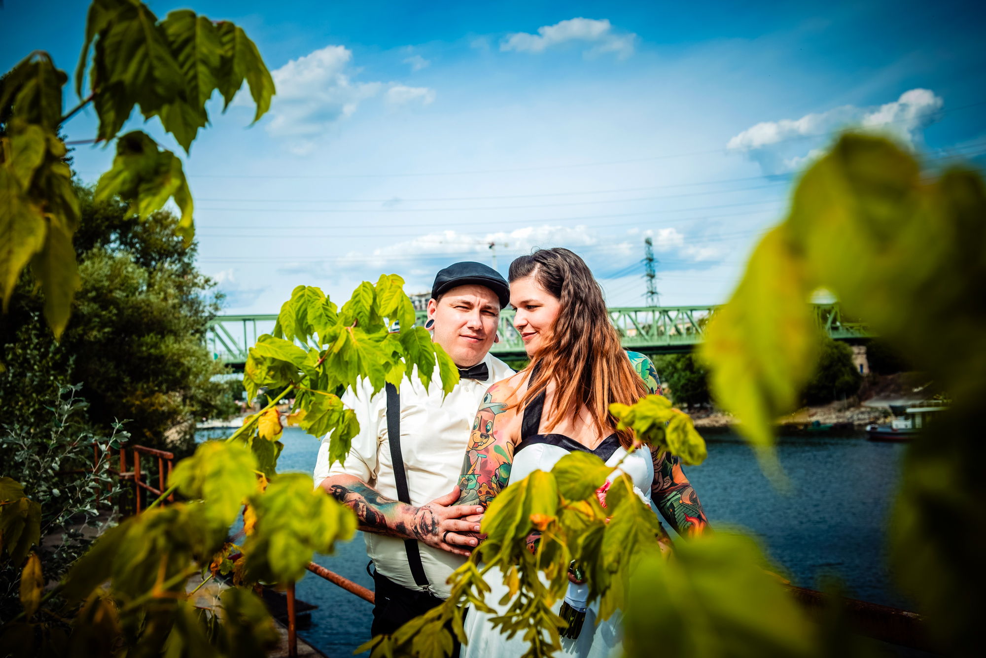 Alternative newlyweds standing beside riverside greenery with bridge in the background near Budapest.