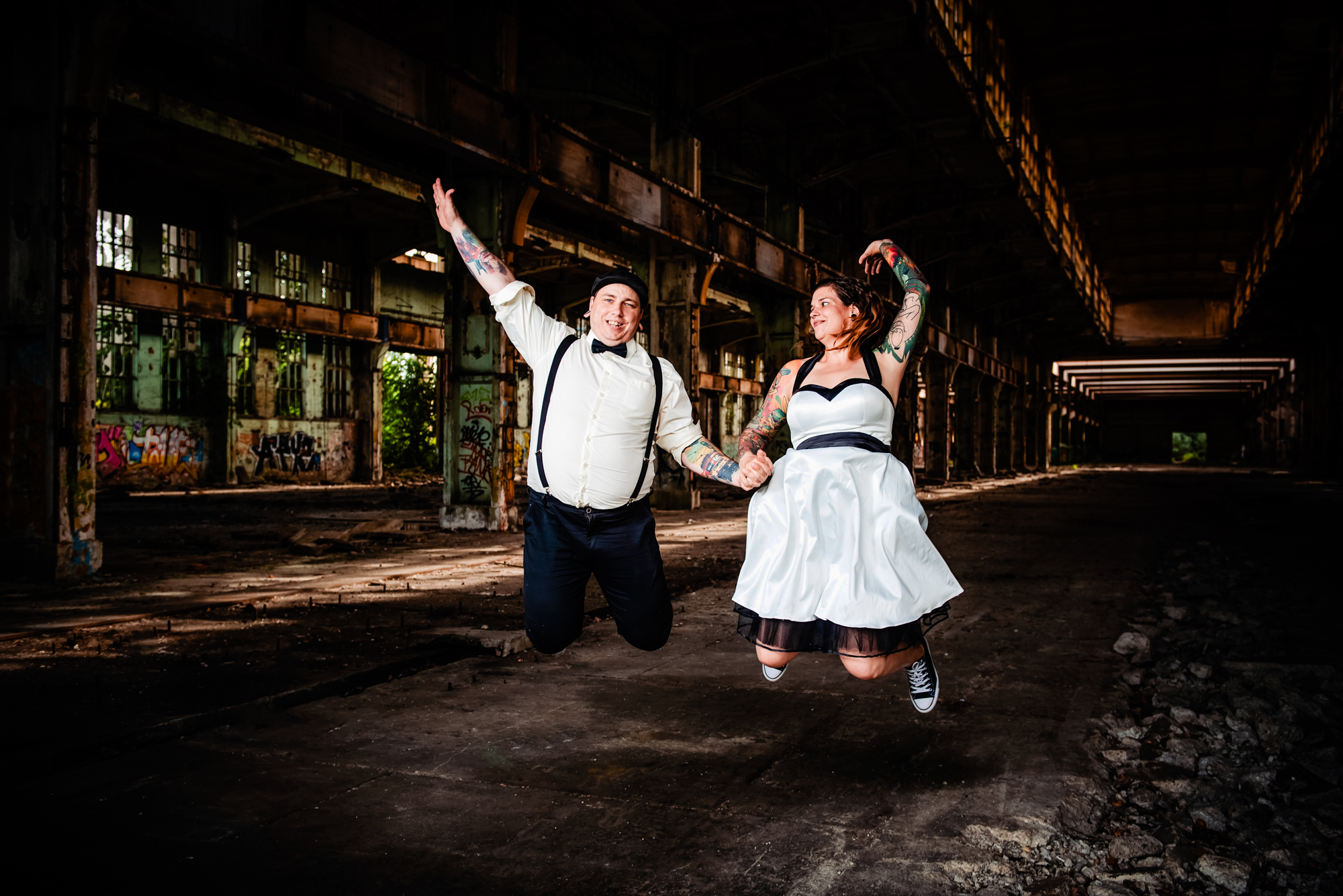 Alternative wedding couple jumping together inside abandoned industrial warehouse during Budapest portrait session.